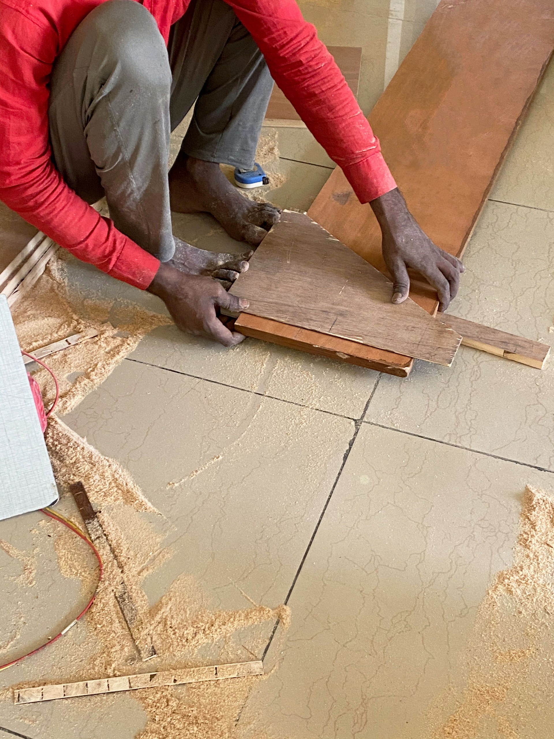 Close-up image of Indian handyman crouching and using off cut of plywood to measure wooden plank, tiled floor covered in sawdust