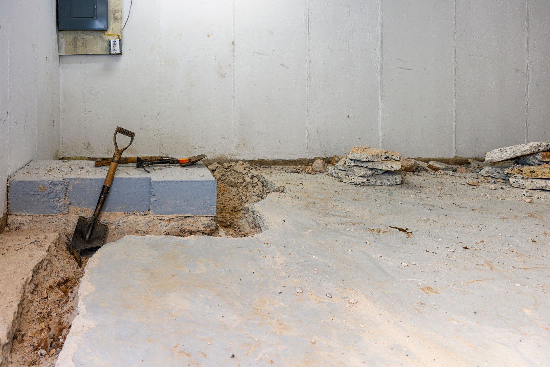 Excavating perimeter trench for drainage solution. Unfinished basement reveals concrete removal work and gray block foundation preparation for water control system